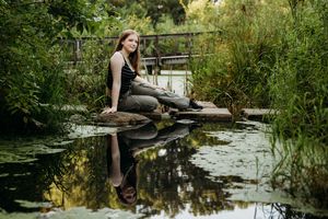 Person in a black dress sits by a peaceful pond surrounded by lush greenery on a sunny day.
