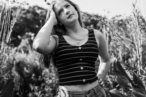 Black and white portrait of a person in a striped tank top and jeans standing among wildflowers in nature.