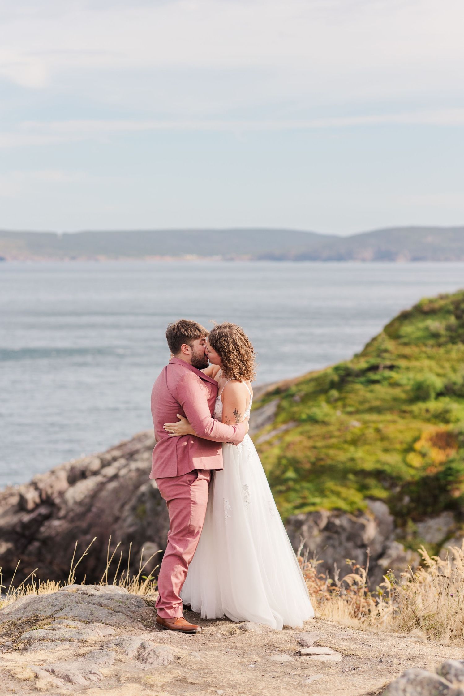 A couple sits together on coastal rocks overlooking the ocean with green cliffs and distant hills in view.
