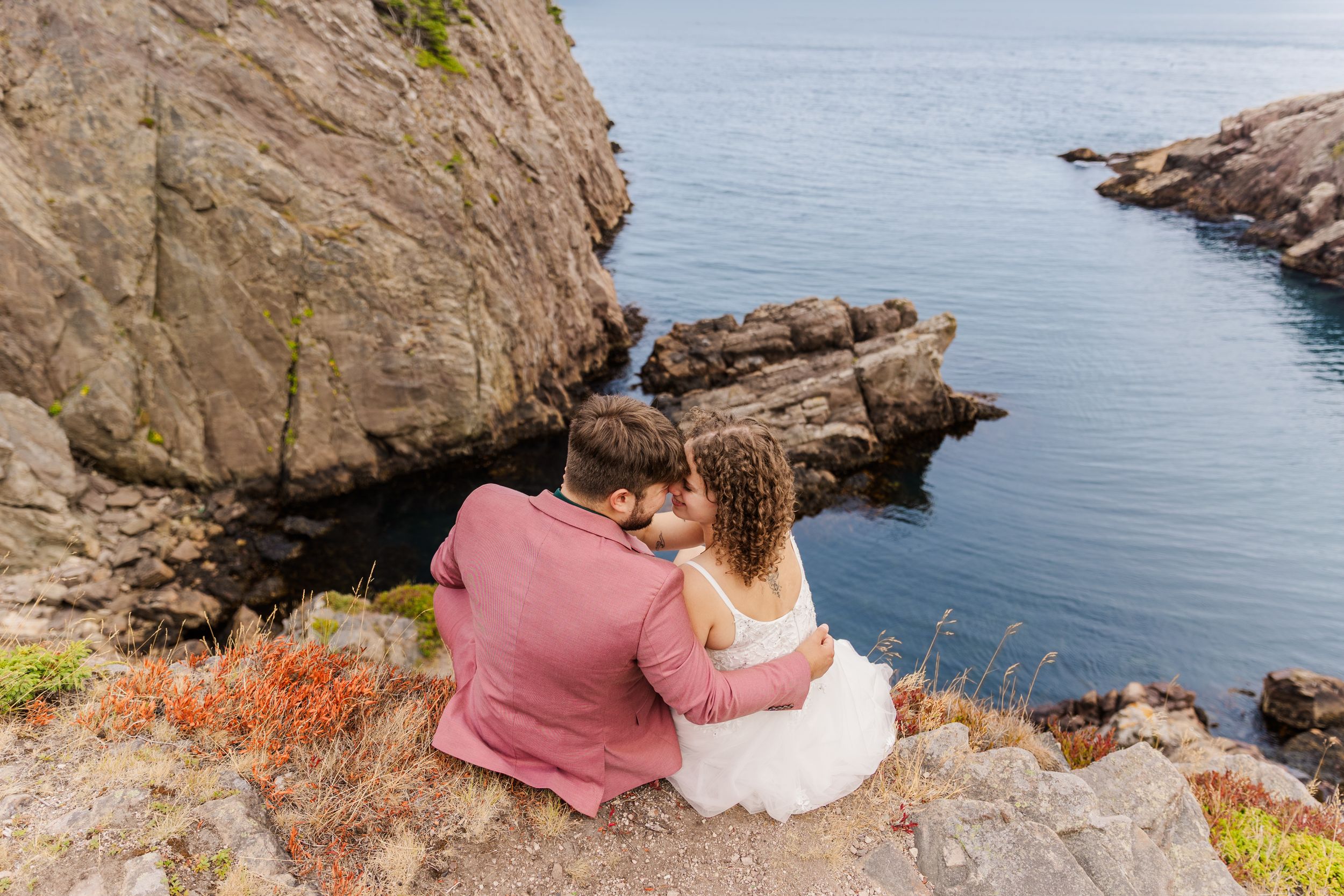 Two people sit on a coastal cliff edge overlooking a pristine blue ocean on a sunny day with rocky outcrops visible.