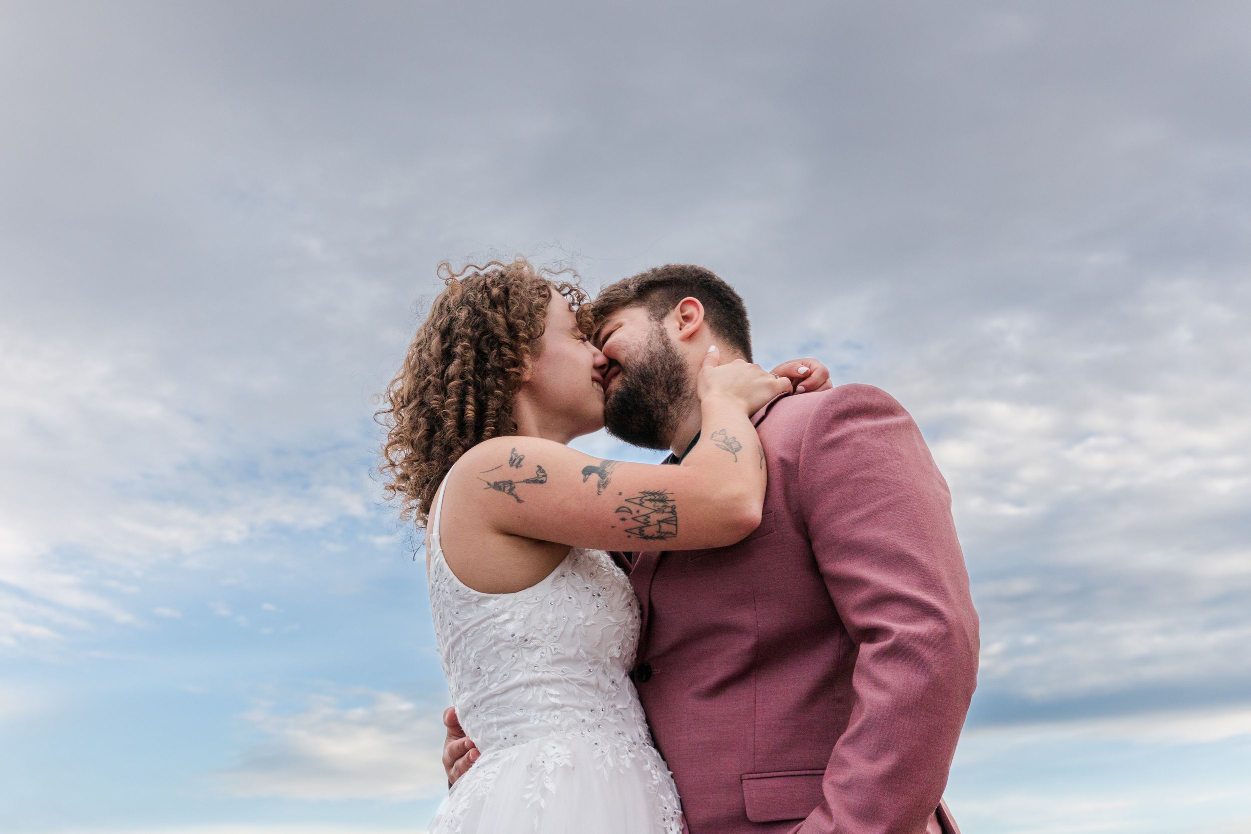 A couple embraces and kisses during a romantic outdoor engagement photoshoot against a cloudy sky.