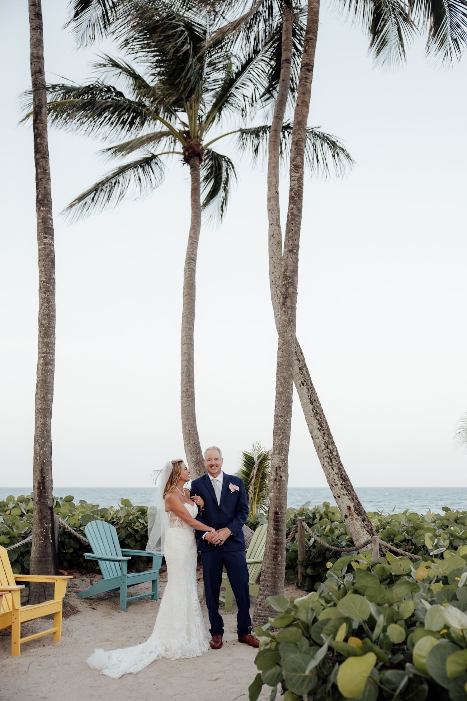 A couple in traditional wedding attire poses for romantic photos on a balcony surrounded by palm trees in a sunny garden setting.