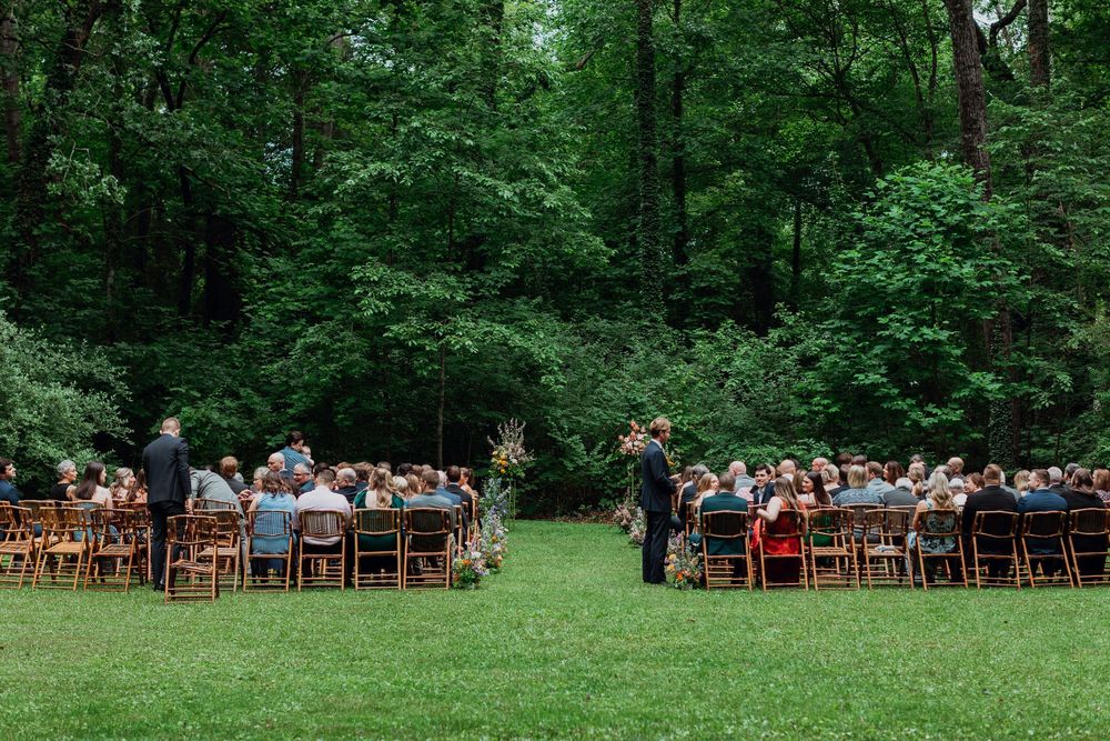 Outdoor wedding ceremony setup with wooden chairs arranged on a lush green lawn surrounded by forest trees.