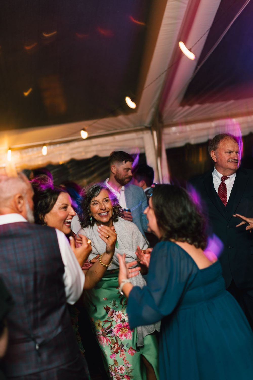 Wedding reception guests dancing and laughing under a tent with colorful lighting.