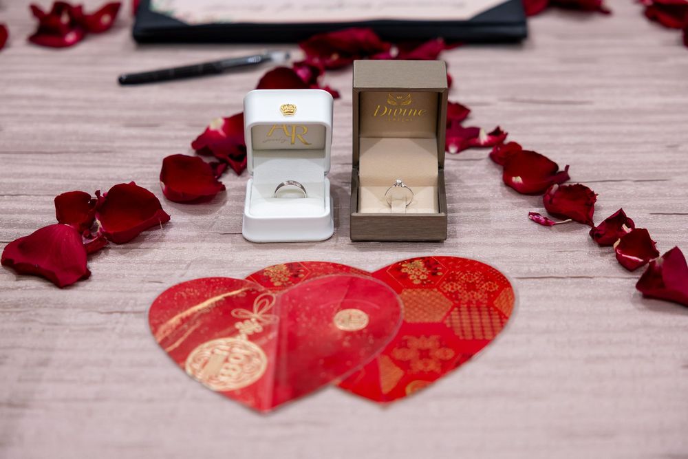 Wedding rings in white boxes with red rose petals and traditional Chinese red envelope decorations on wooden surface.