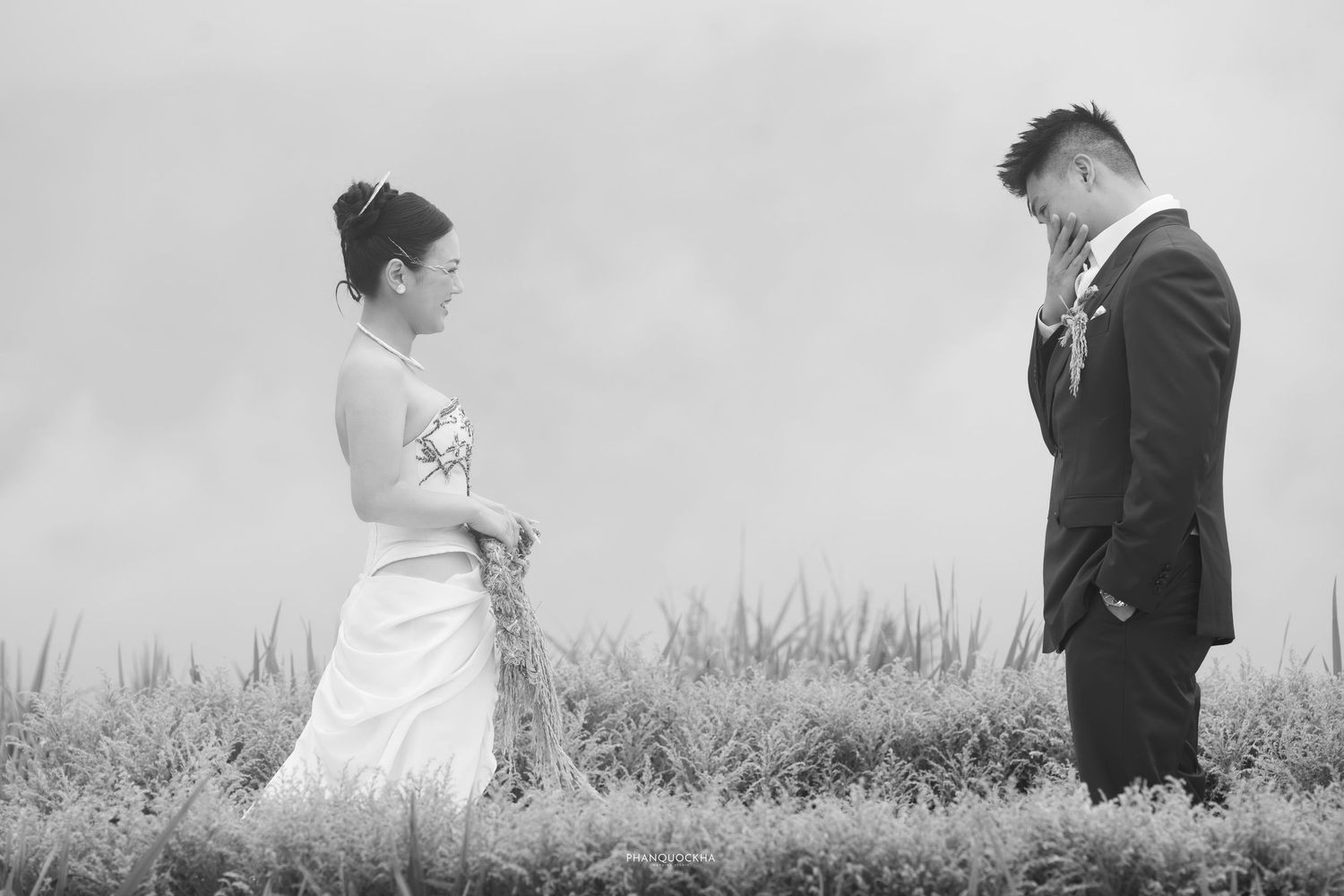 Black and white wedding portrait of Asian couple standing apart in tall grass field, groom bowing his head.