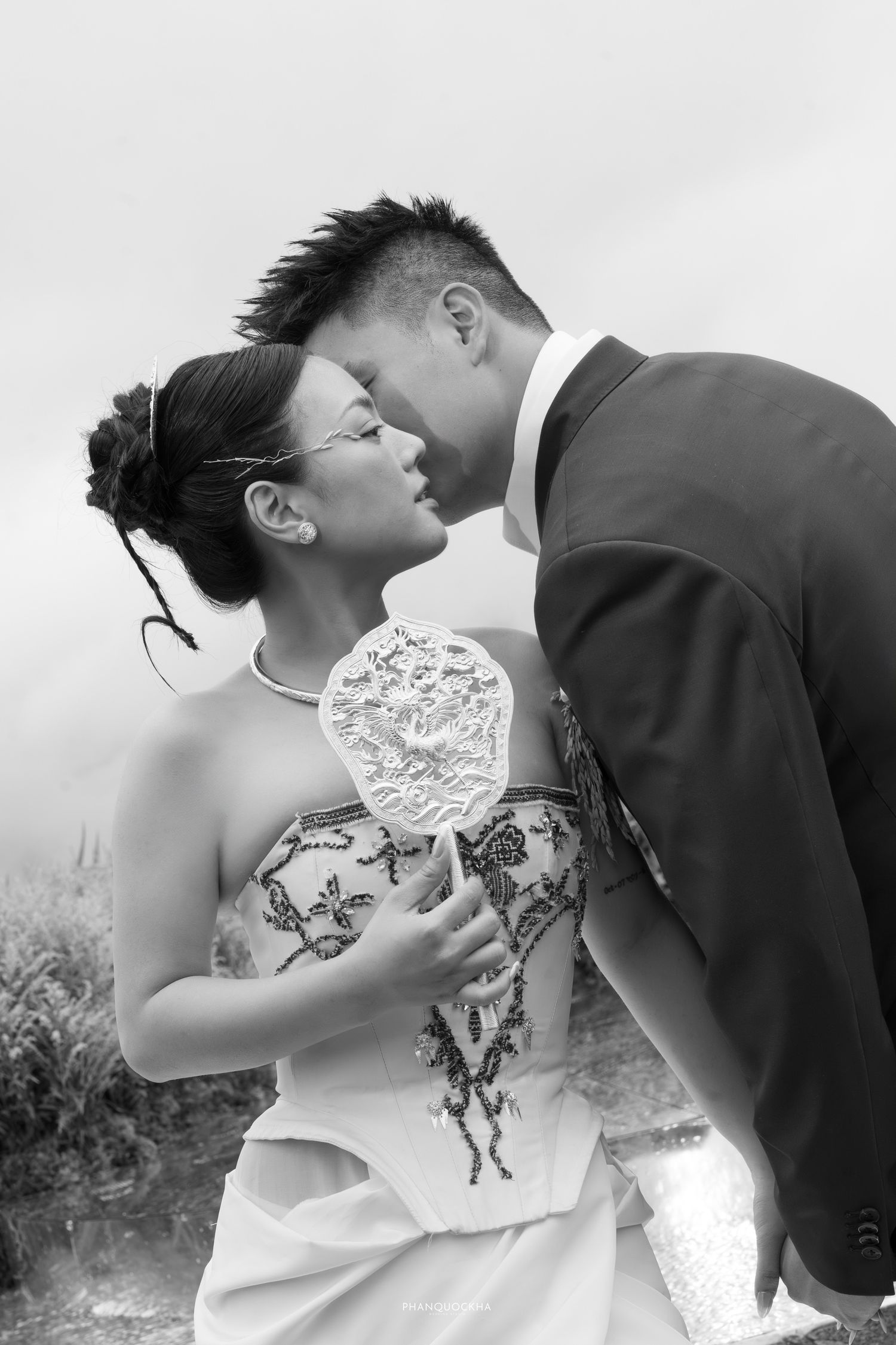 Black and white photo of Asian wedding couple sharing a tender kiss, bride holding lace fan in flowing strapless gown.