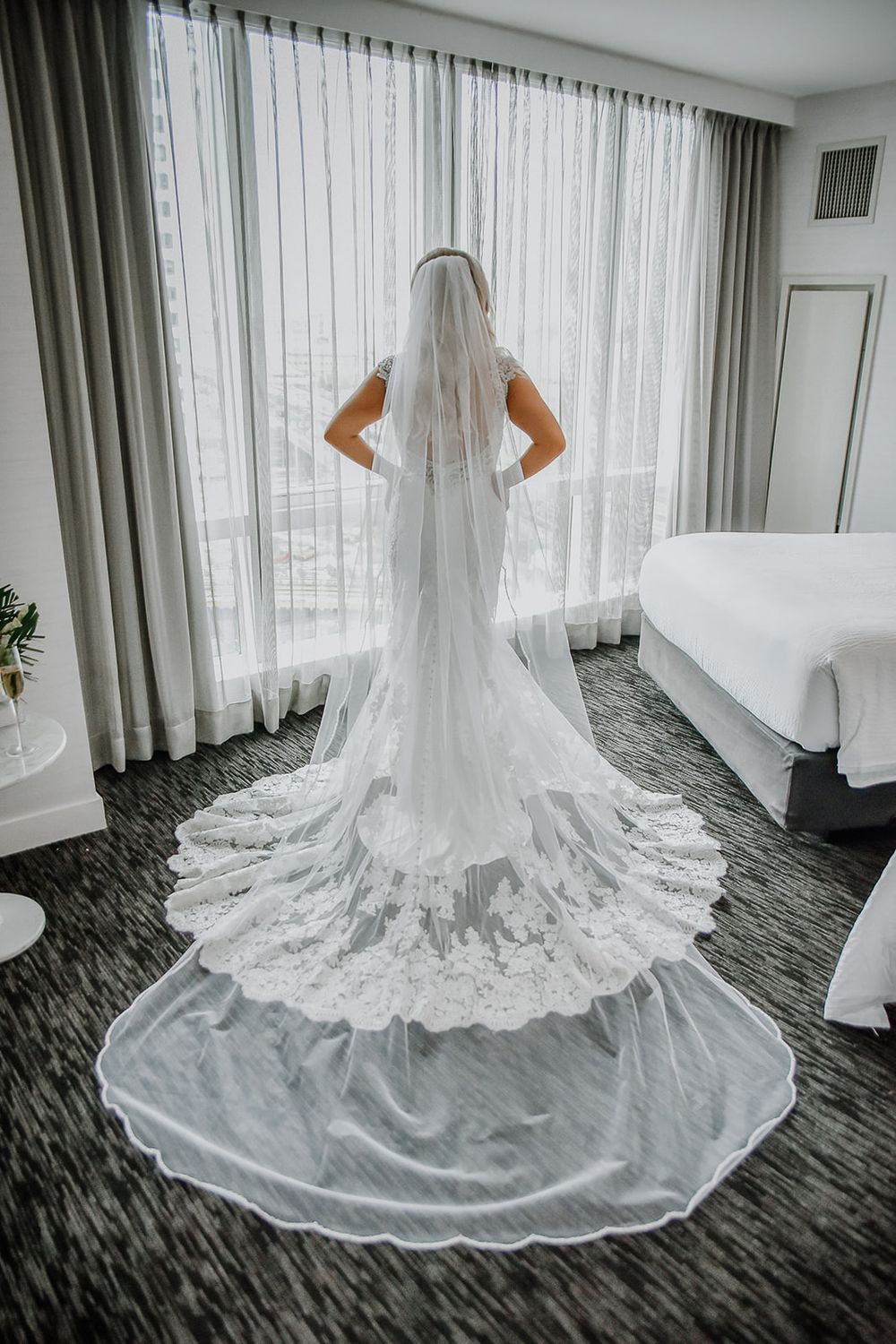 A stunning white lace wedding dress with cathedral length train displayed on dark patterned carpet near large windows.