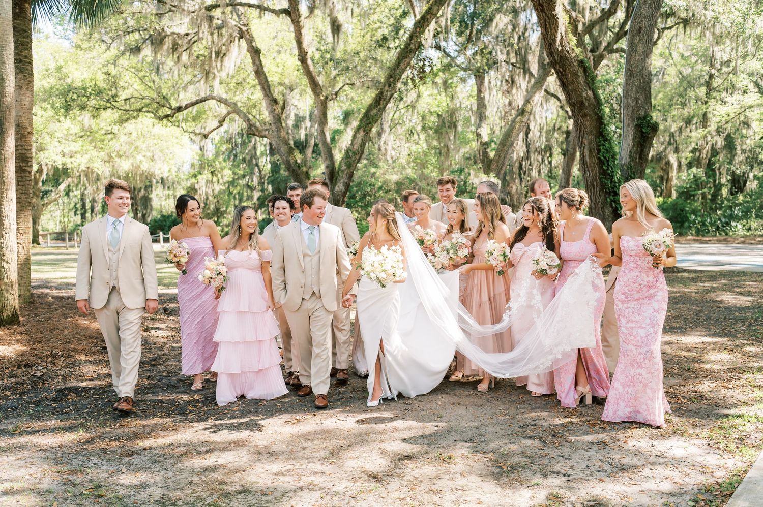 Wedding party dressed in pink and white dances and celebrates joyfully along a tree-lined path on a sunny day.