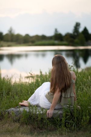 Figure in a white dress sits peacefully by a lake at sunset, surrounded by tall grass and natural scenery.