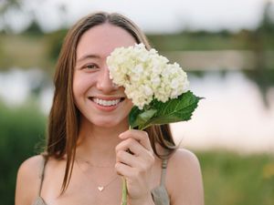 A white hydrangea bloom is held up next to a bright smile outdoors by a lake on a summer day.