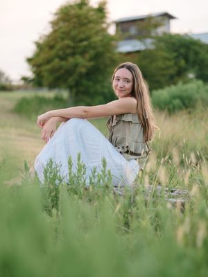 Someone wearing a casual olive top and white pants sits in tall summer grass during golden hour portrait photography.