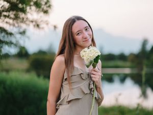 Woman in a beige dress holds white hydrangea flowers by a serene lake at dusk.