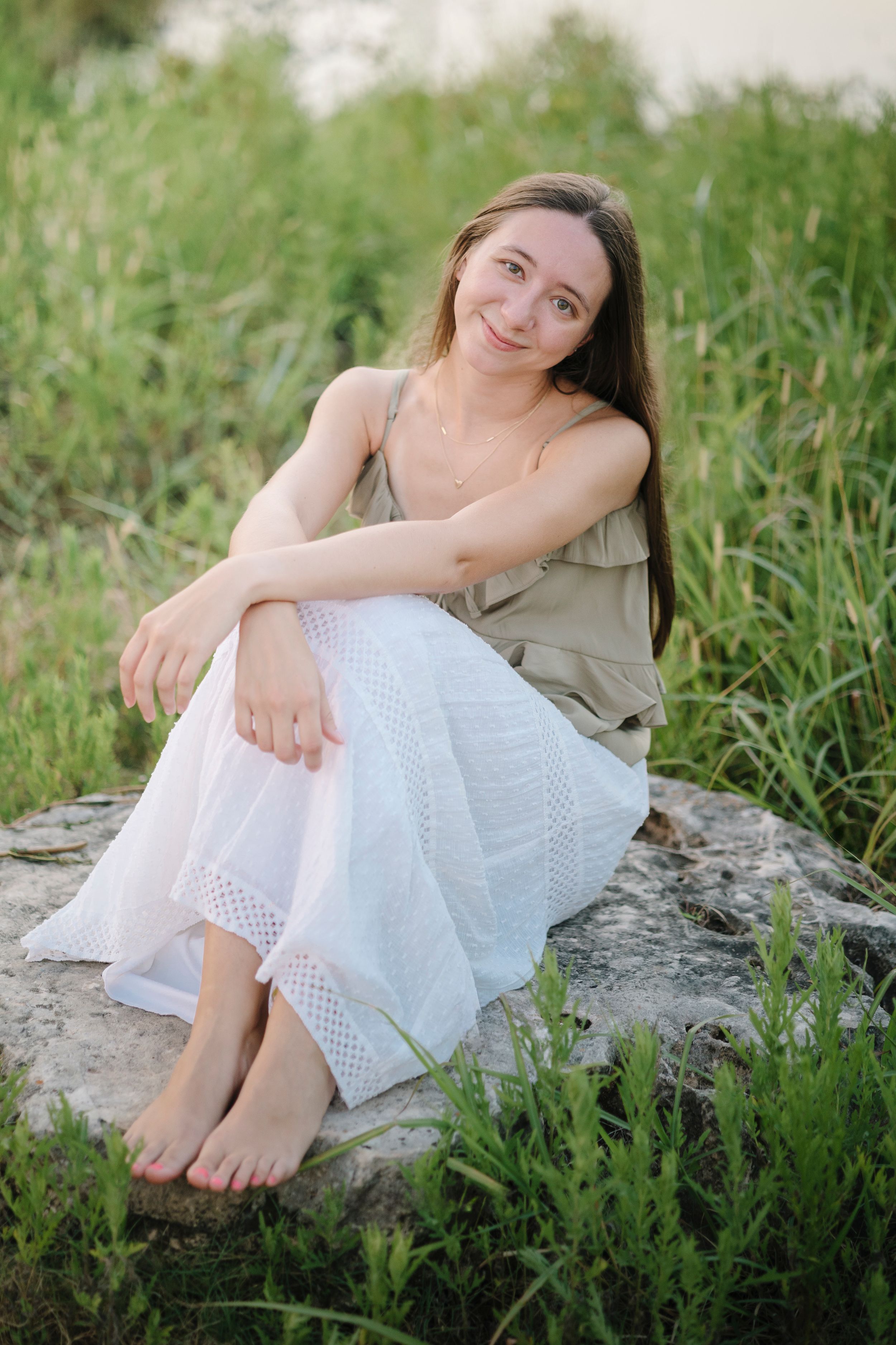 Person in sage dress and white skirt sits on rock amid greenery by waterside in warm natural lighting.