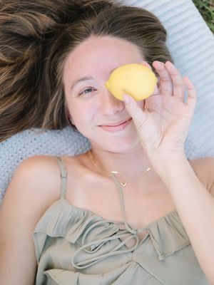 Someone lying down on a white surface holds a fresh yellow lemon in front of their eye while smiling softly.