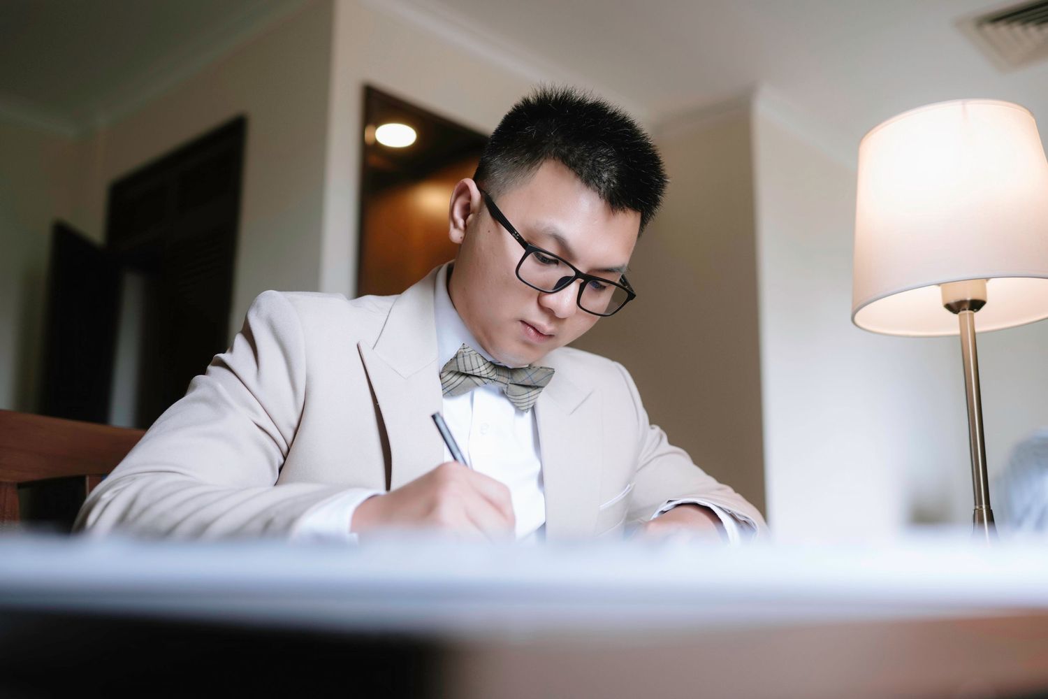 Asian groom in beige suit and bow tie writing at a desk by a lamp.