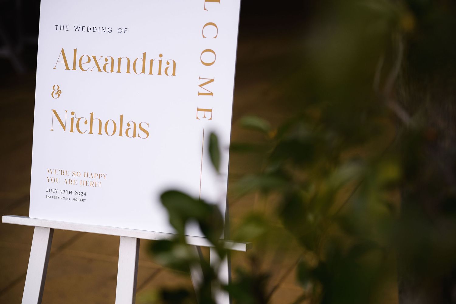 Elegant white wedding ceremony signs with gold text displaying names and details stand in soft focus.