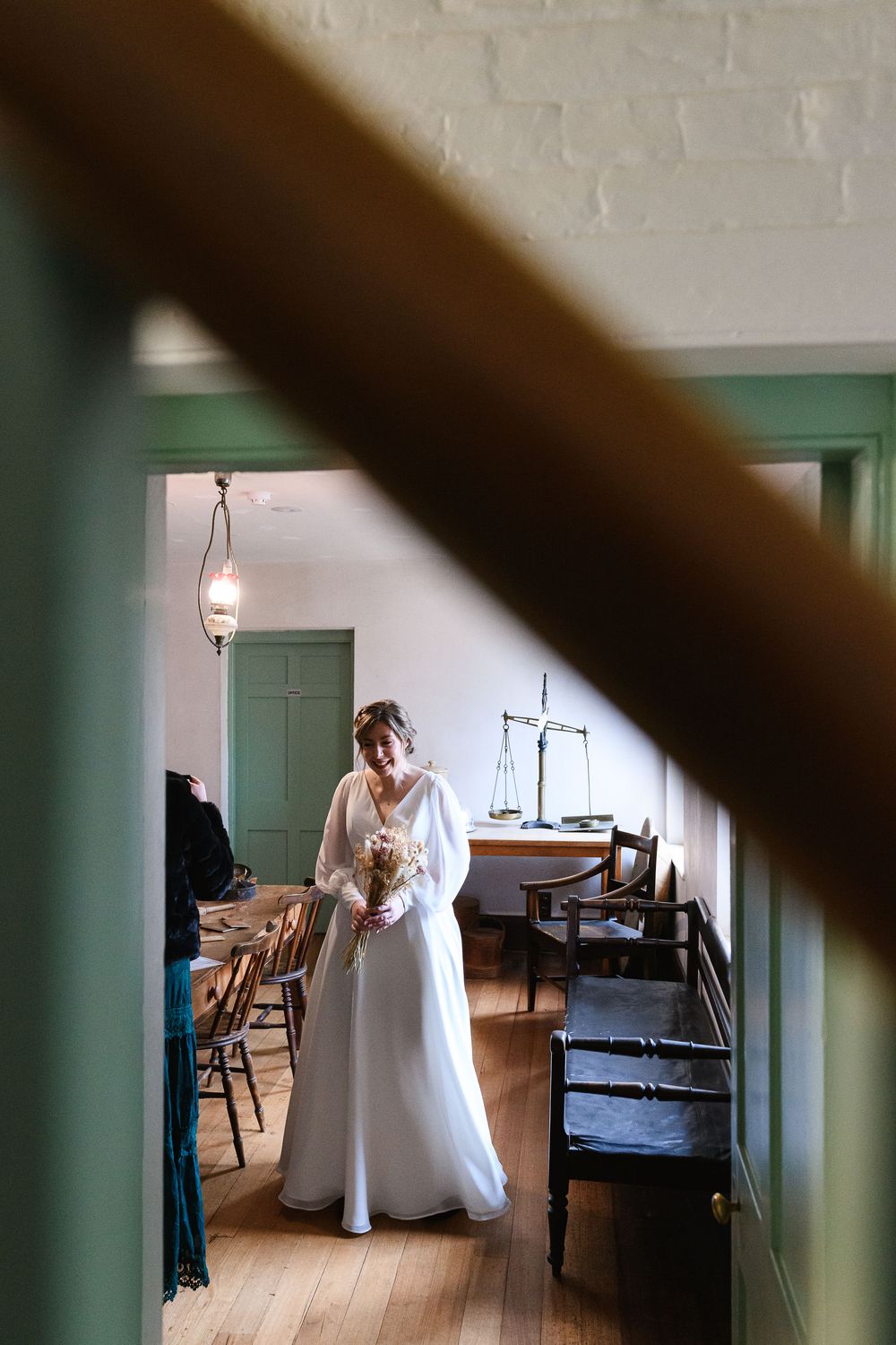 A person wearing a flowing white wedding dress stands in a vintage room with wooden floors and green trim.