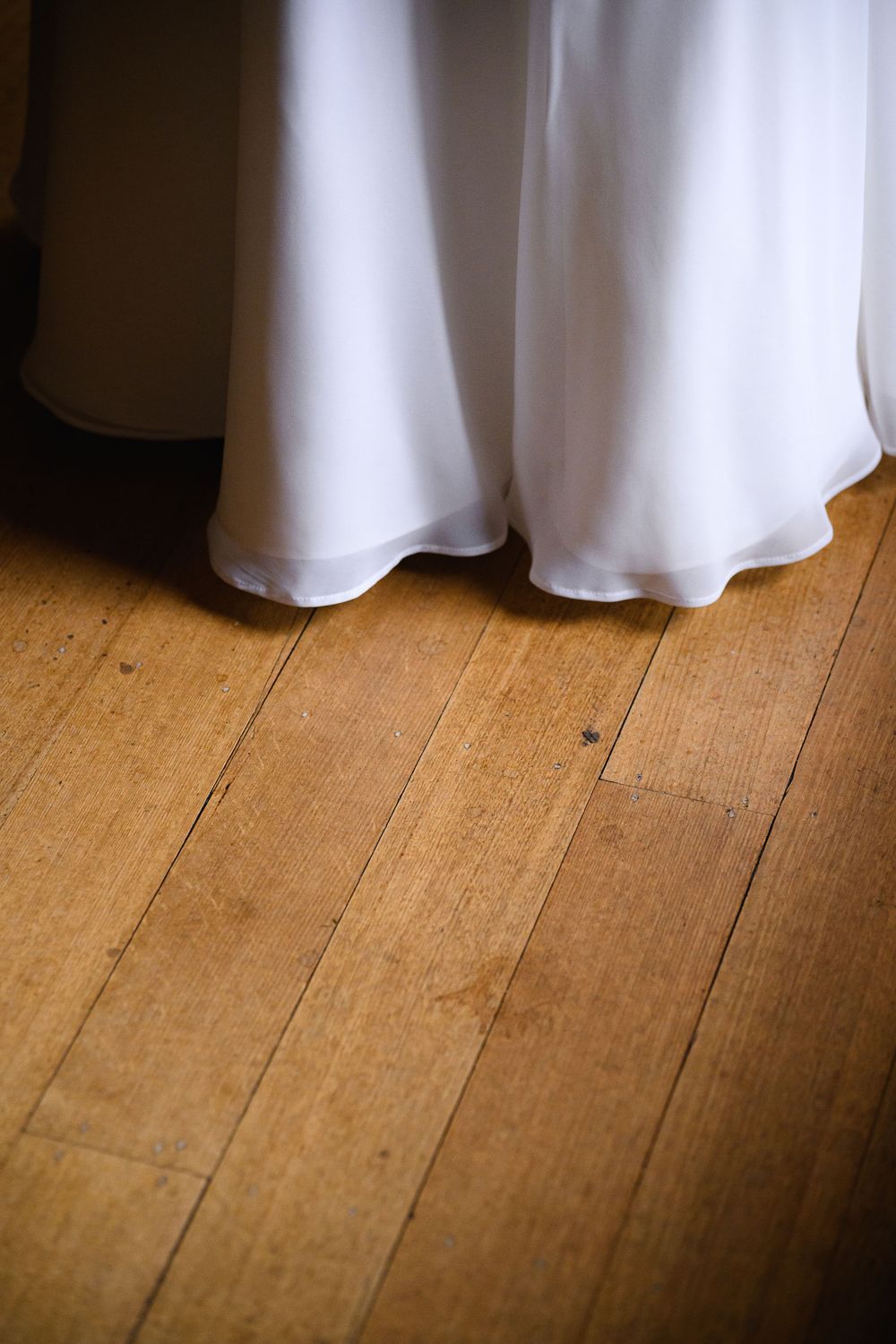The hem of a white wedding dress flows gracefully over rustic hardwood floorboards in warm lighting.