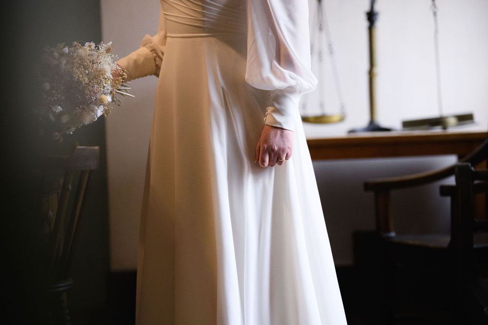 A flowing white dress catches light near a window, creating an ethereal bridal portrait with soft natural lighting.