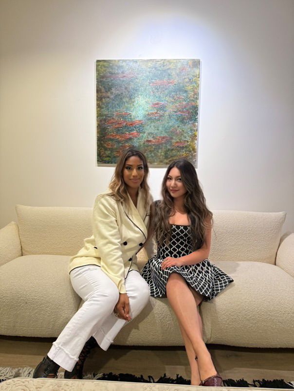 Modern art decor adorns the wall above a cream-colored sofa where two friends sit together in contrasting outfits.