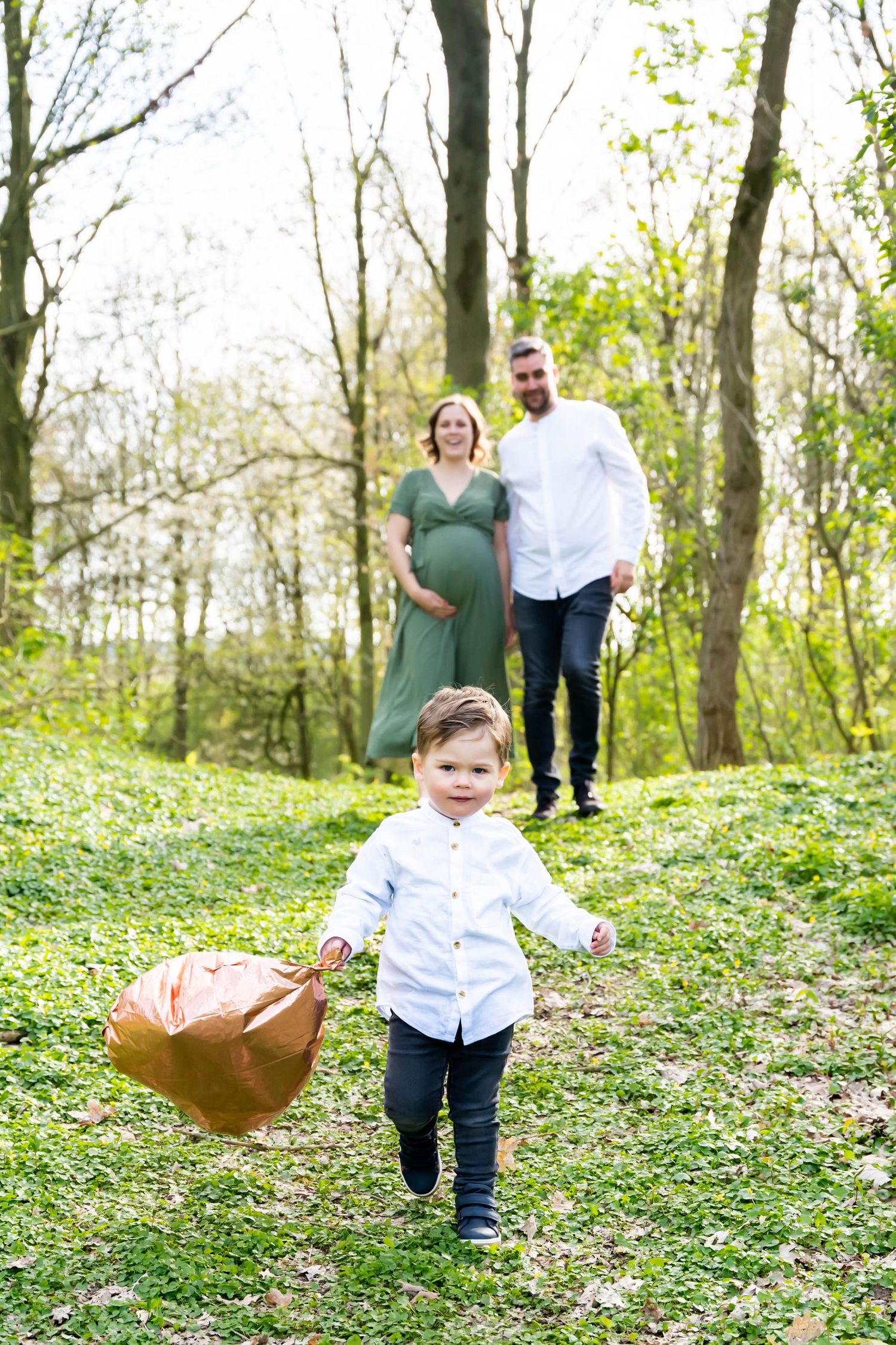 Sweet family moment outdoors with maternity shoot in wooded area as toddler carries brown balloon on grassy hill.