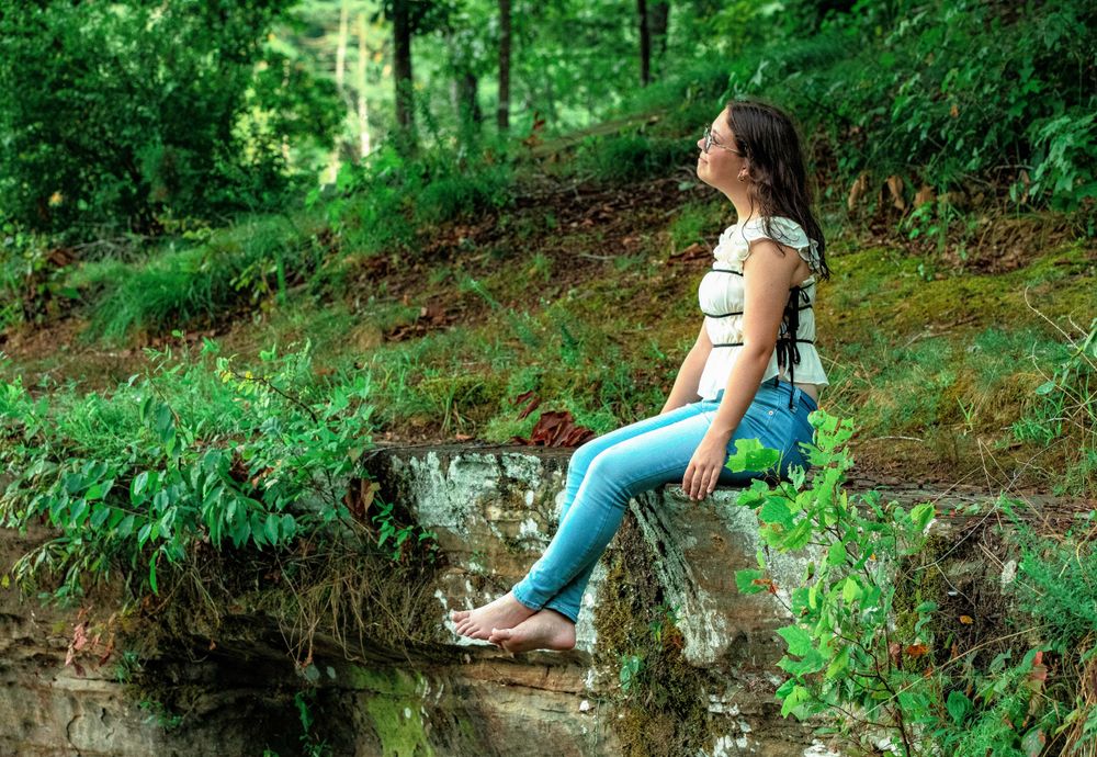 Someone sits casually at the edge of a creek bank surrounded by lush greenery.