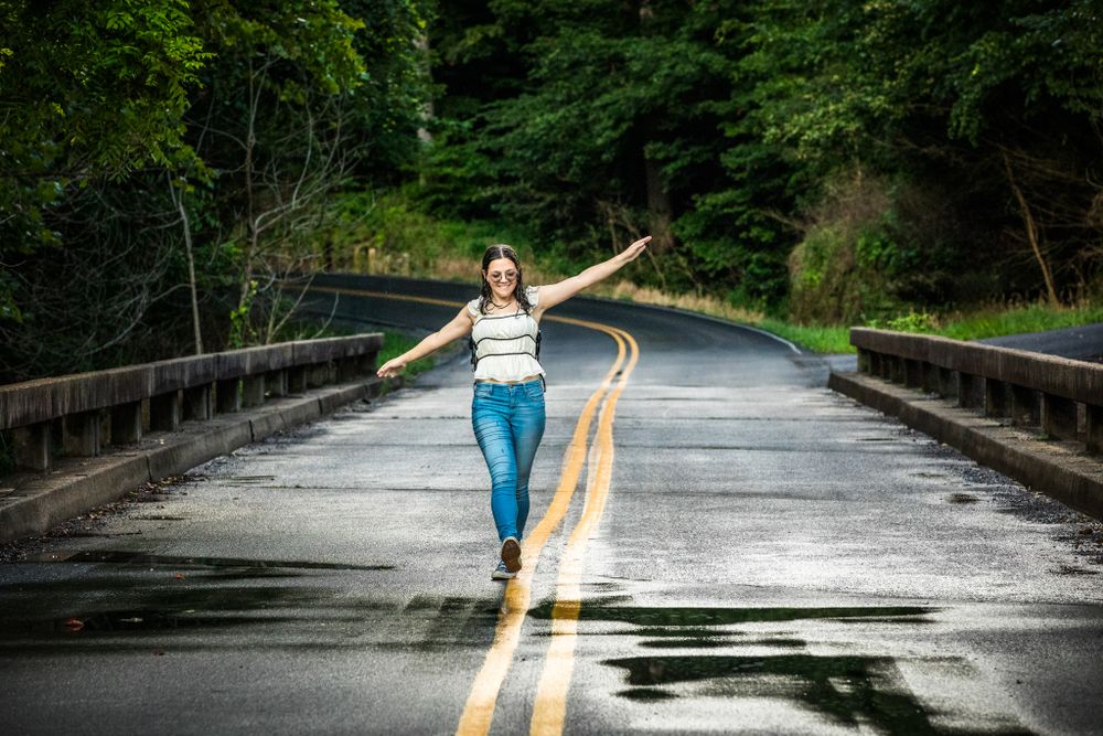 A person in casual attire balances on yellow road lines of a wet bridge surrounded by forest.