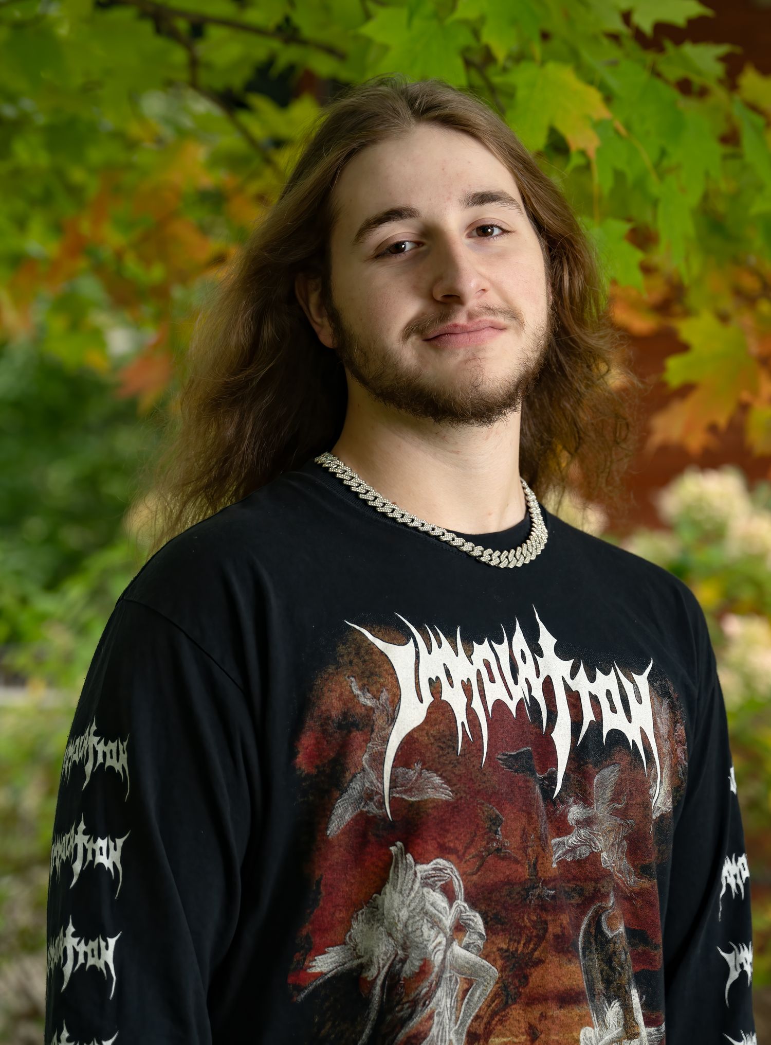 Young person in black metal band shirt with long hair posing against autumn foliage.