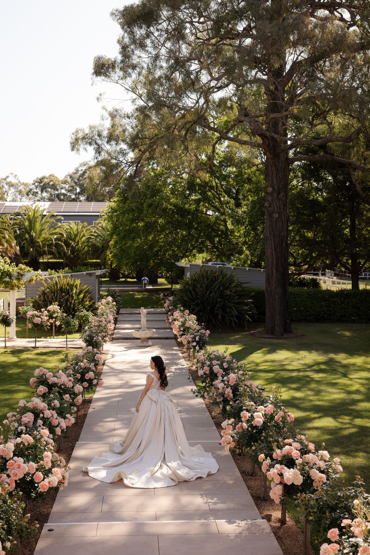 Bride in ball gown walking along flower-lined aisle path in lush garden wedding venue.