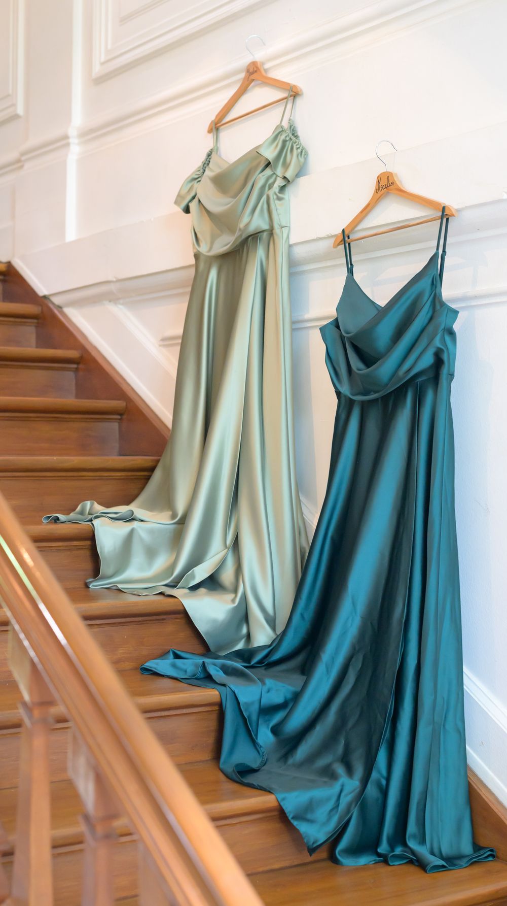 Two elegant bridesmaid dresses in mint and teal colors hanging on wooden hangers against a white wall with staircase.