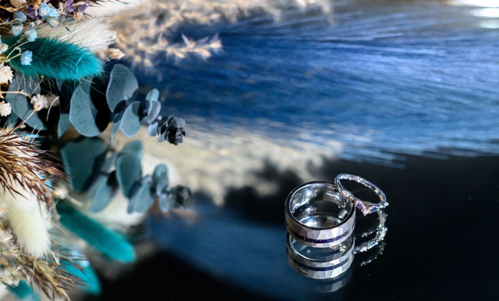 Wedding rings captured in artistic detail with teal florals and reflective surface in background.
