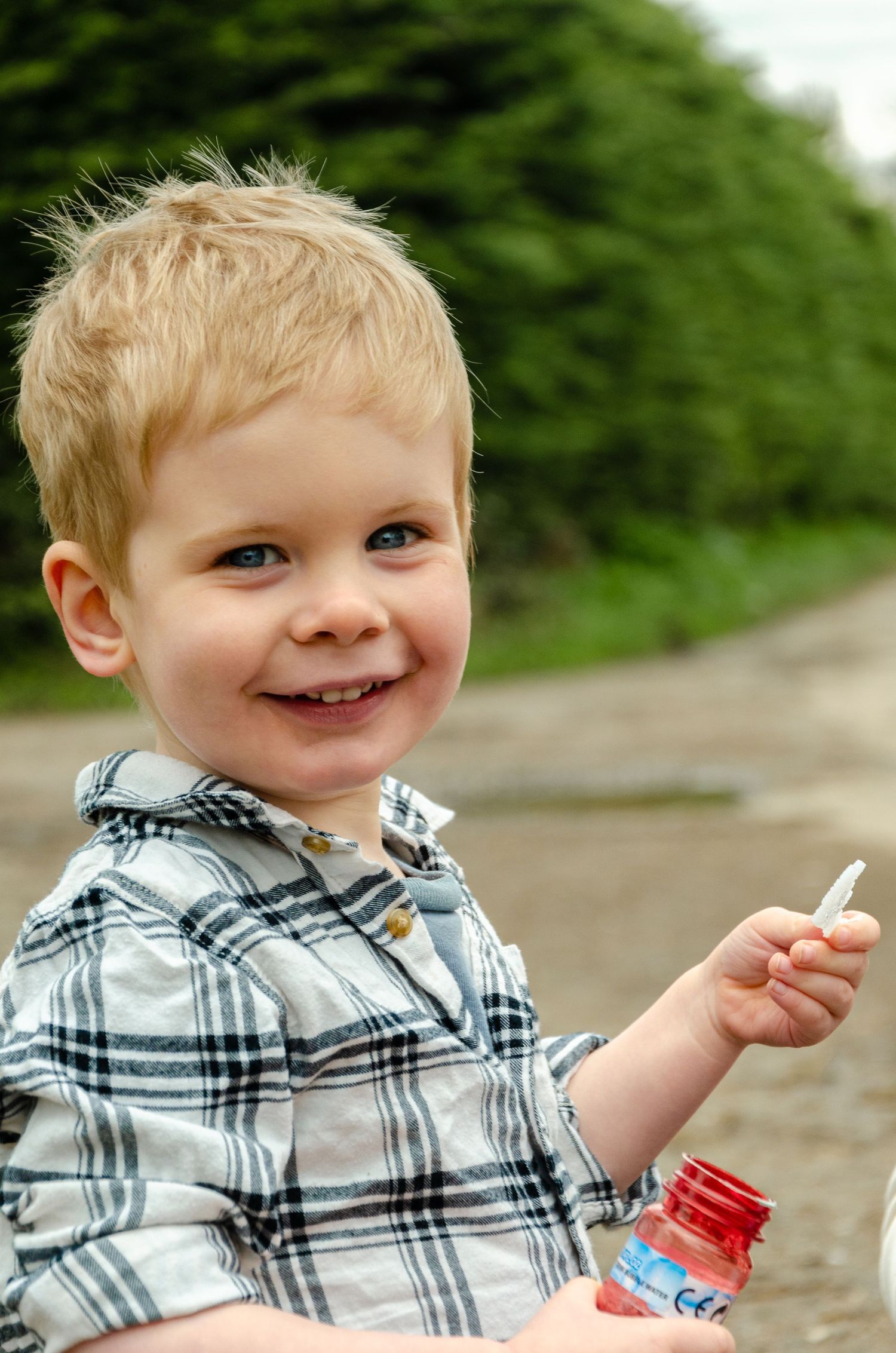 Young child in plaid shirt smiling while blowing bubbles outdoors.