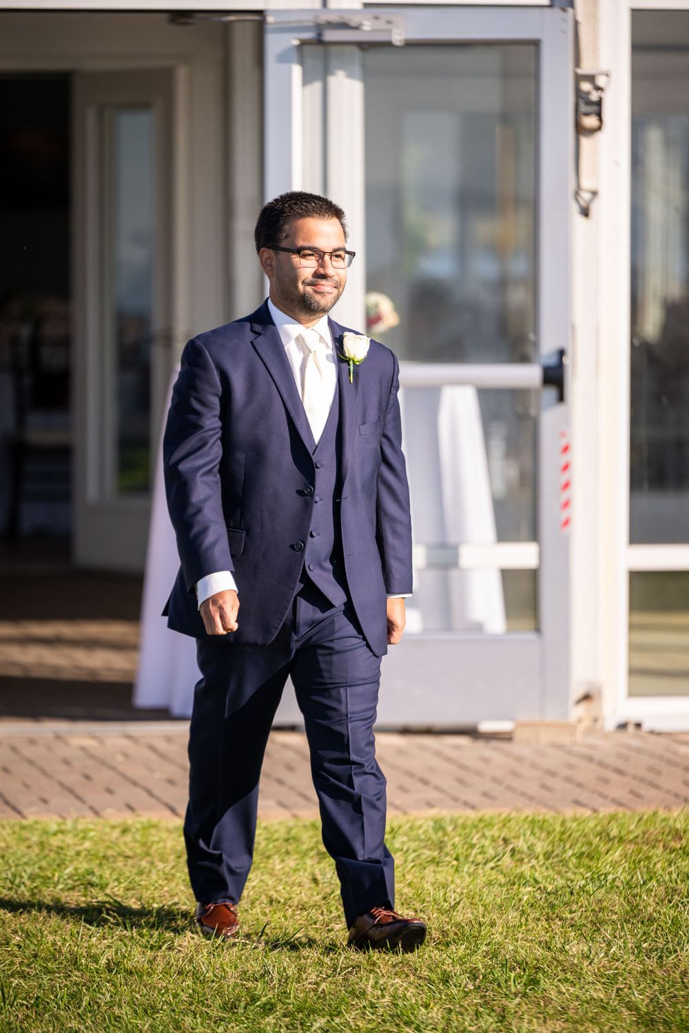 A groom in a navy blue three-piece suit stands on grass outside a venue entrance.