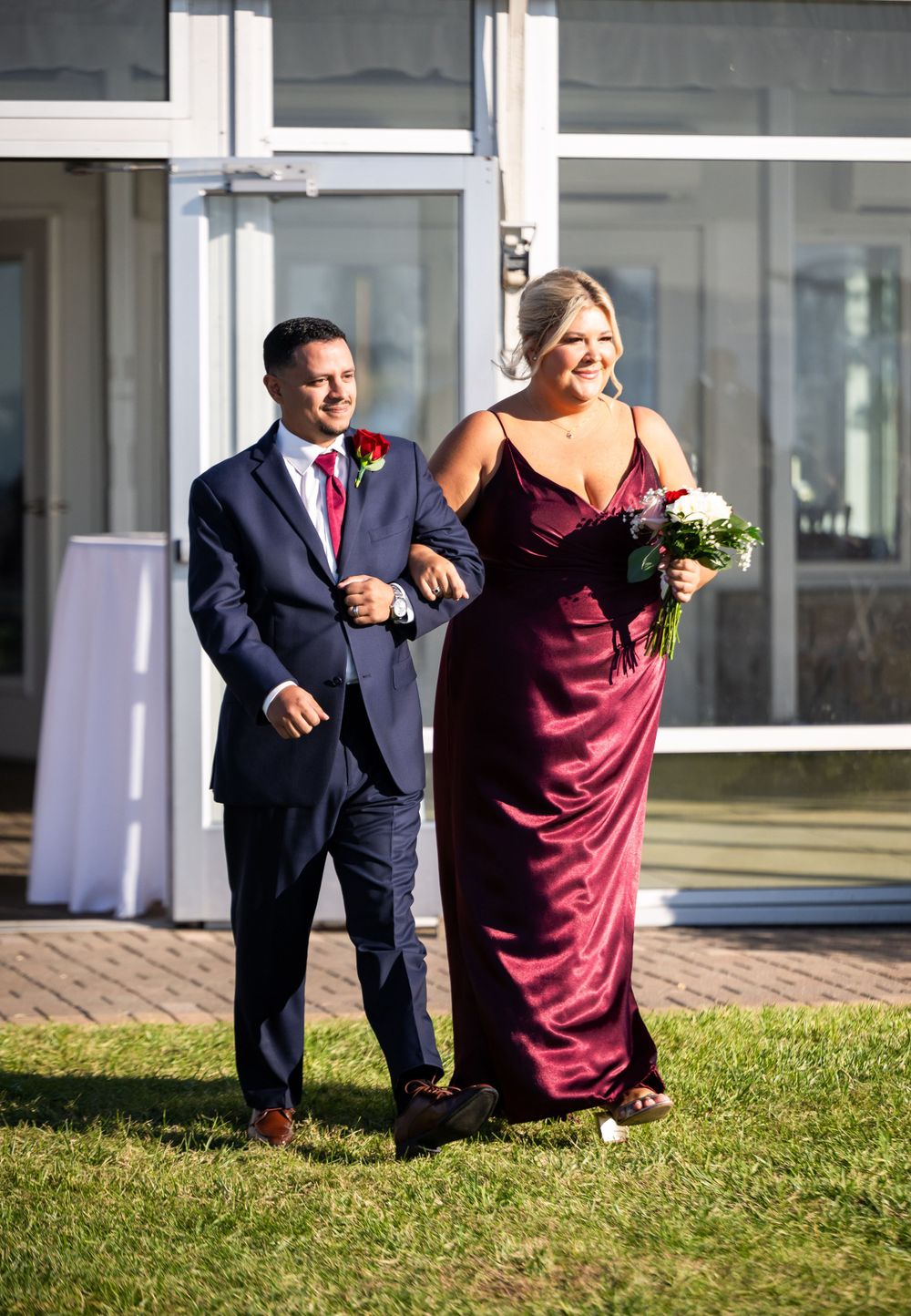 Wedding party members in navy suit and burgundy dress walk arm-in-arm on a sunny path.