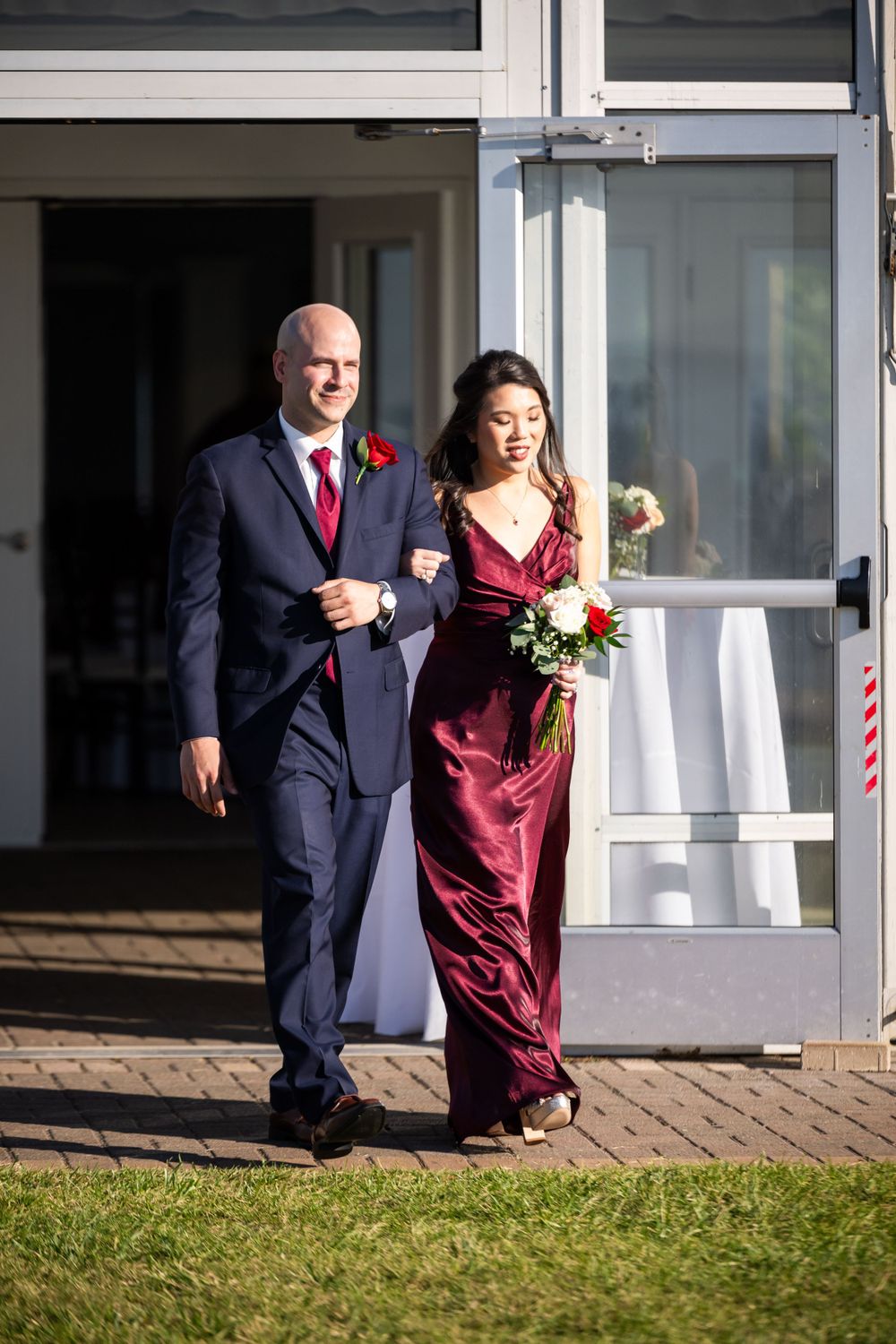 Wedding party members in navy suits and burgundy dresses walking together on brick pathway.