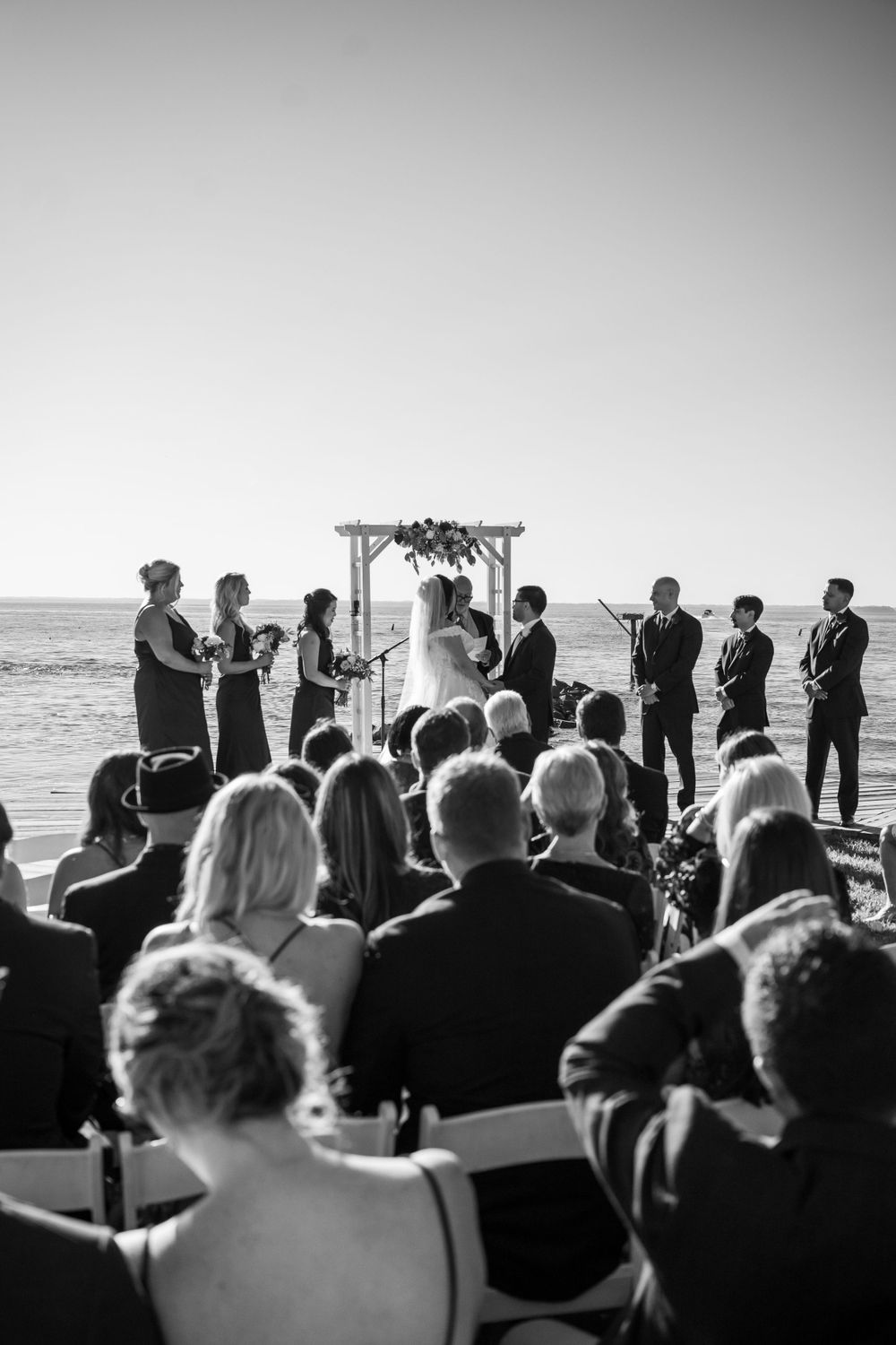 Silhouetted wedding guests watch beachside ceremony under wooden arch.