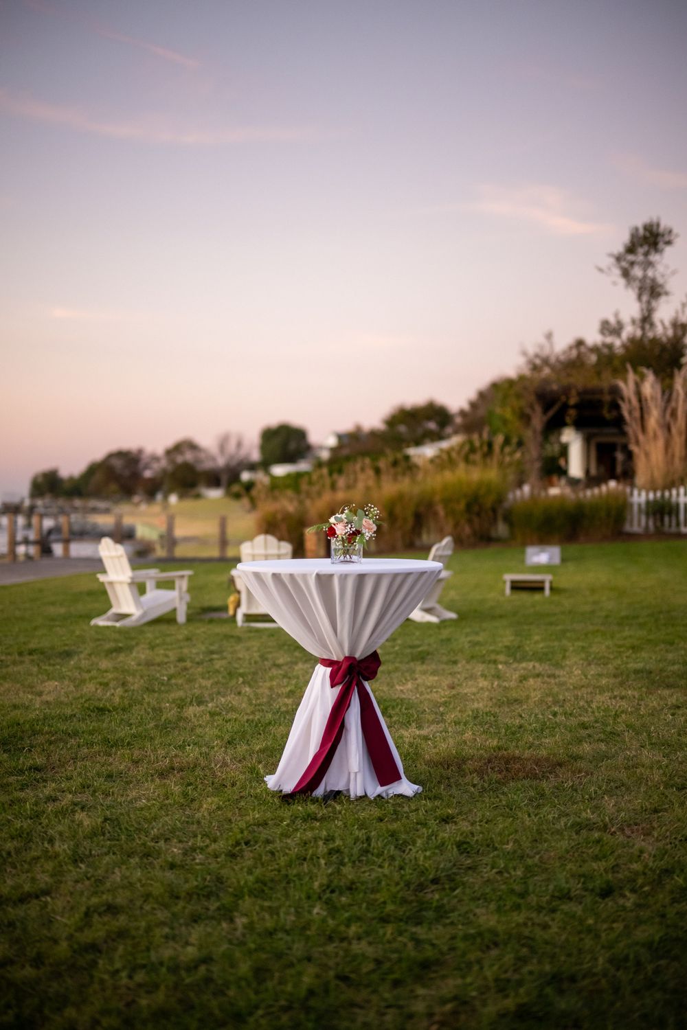 White cocktail table with burgundy sash on waterfront lawn with Adirondack chairs.