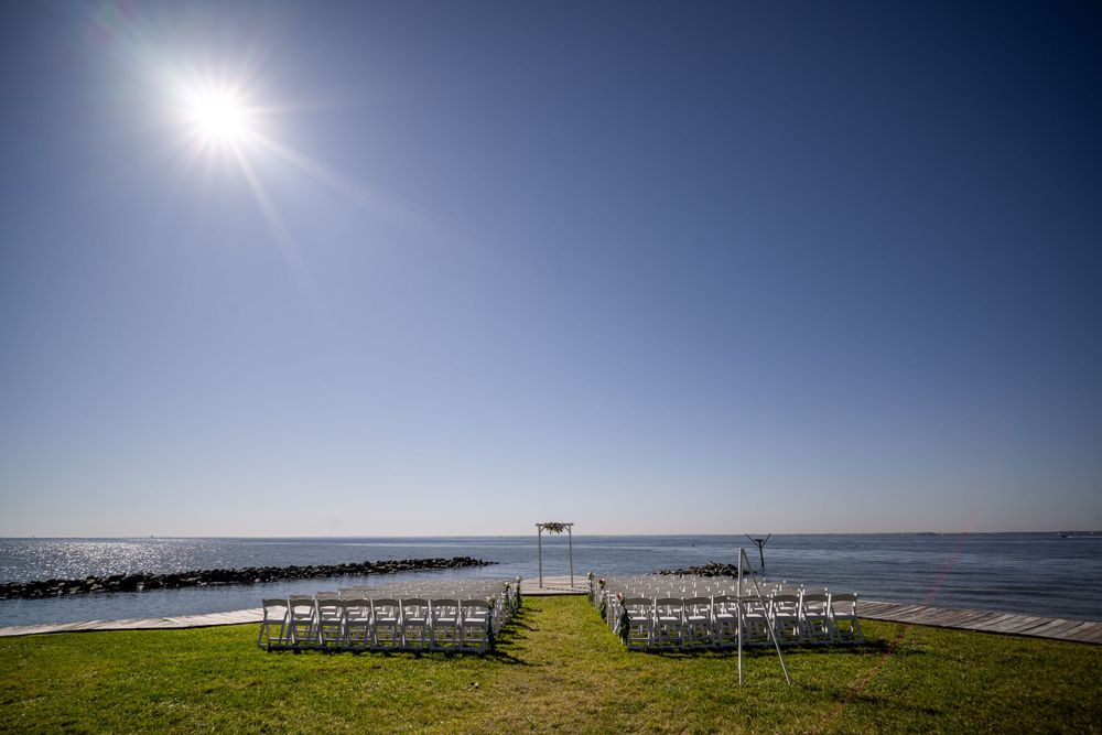 Outdoor wedding ceremony setup on oceanfront lawn with white chairs under bright blue sky.