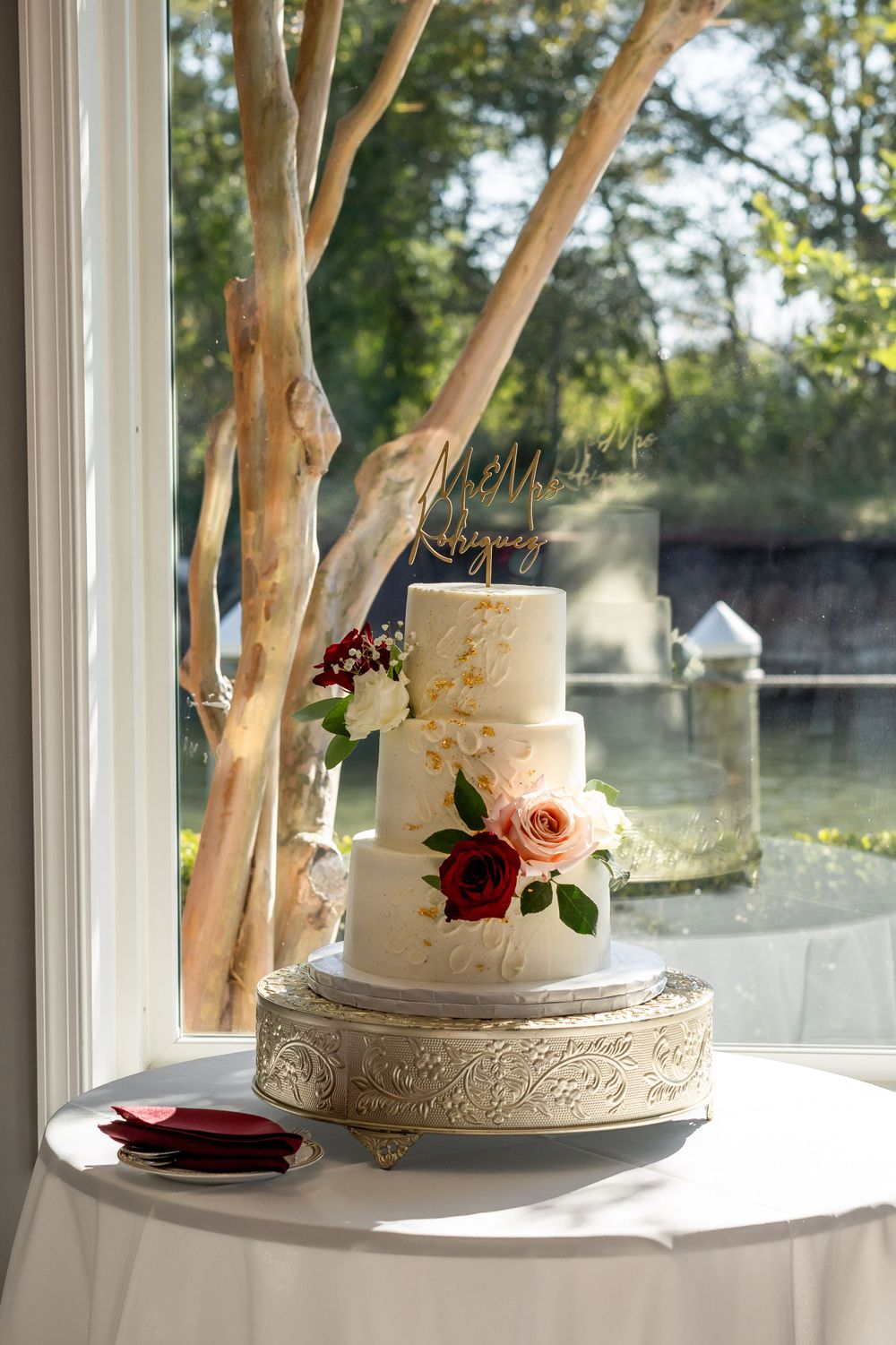 Elegant three-tier white wedding cake with burgundy floral accents displayed on an ornate gold stand by a window.