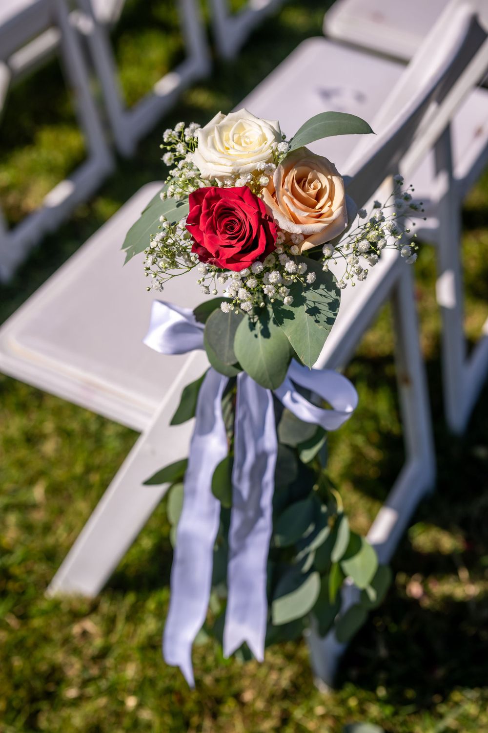 A floral decoration with red and white roses tied with purple ribbon adorns a white ceremony chair.