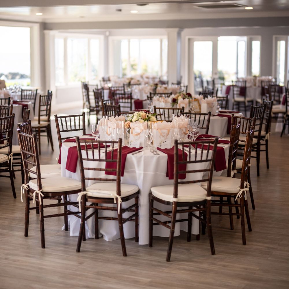 Reception hall set with round tables, burgundy accents, and dark wooden Chiavari chairs.