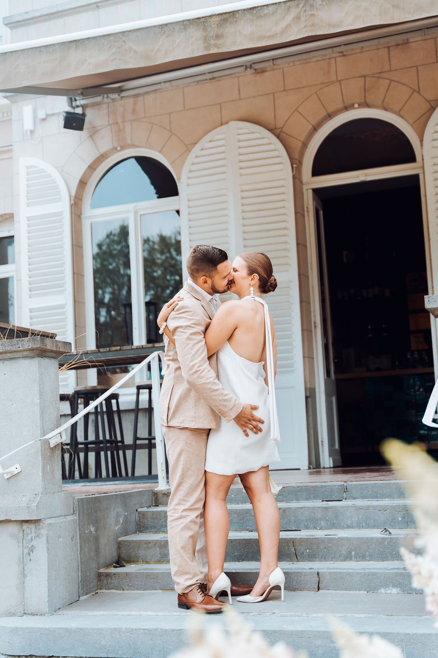 Couple in white dress and beige suit embrace on stone steps by arched windows.