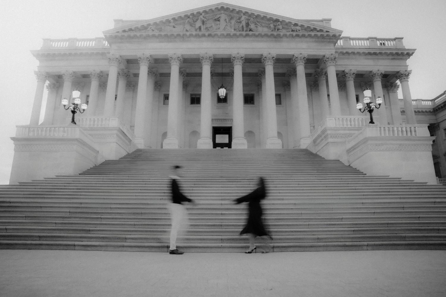 Two blurred figures stand on the steps of a grand neoclassical government building with columned facade in black and white.