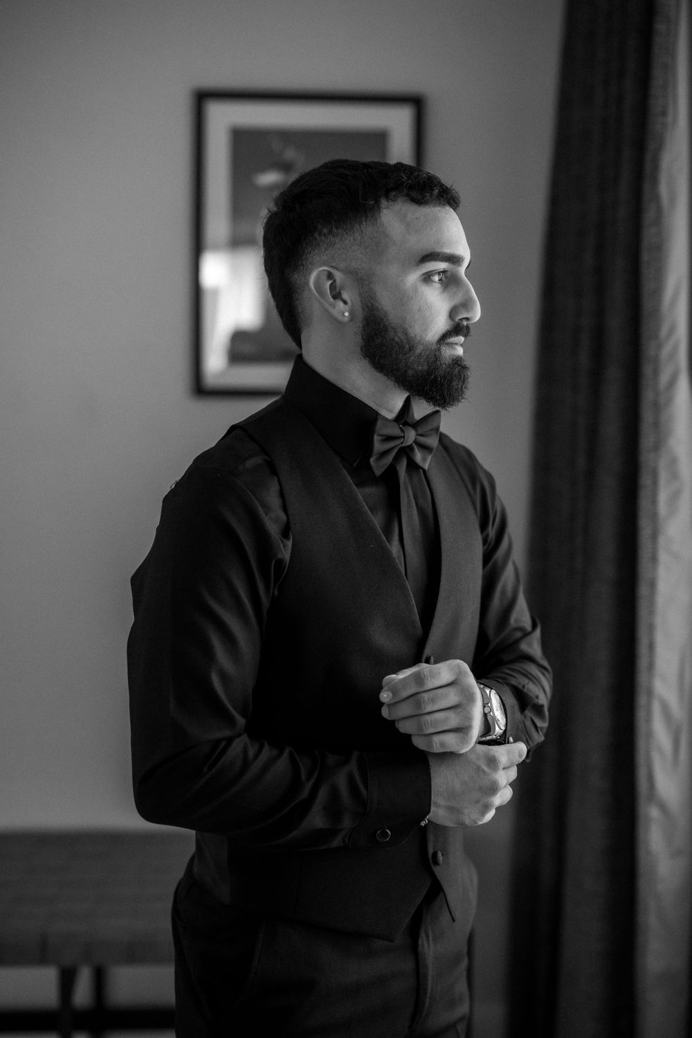 Groom in black tuxedo and bow tie stands pensively by window in monochrome portrait.