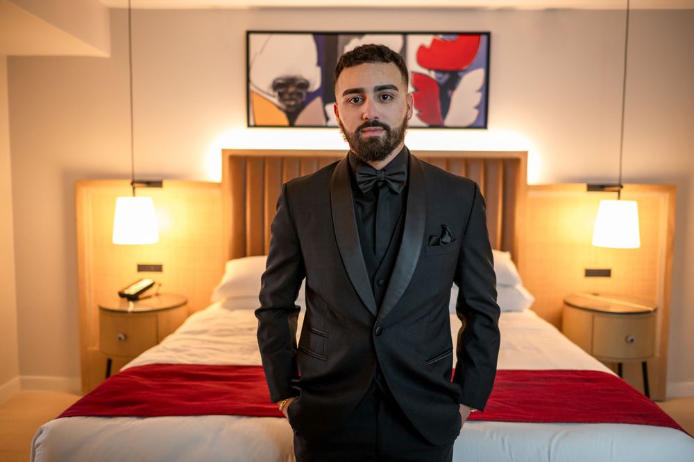 Groom in tailored black tuxedo poses in hotel room with artistic wall decor.