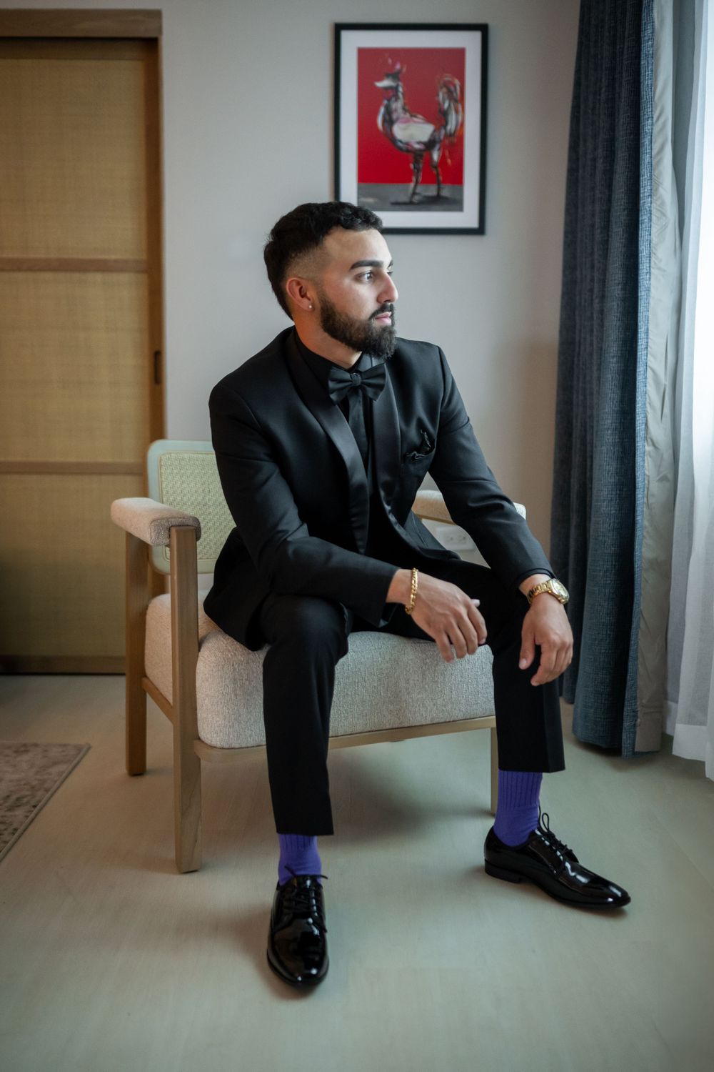Stylish groom in black suit with purple socks sits thoughtfully in modern room.