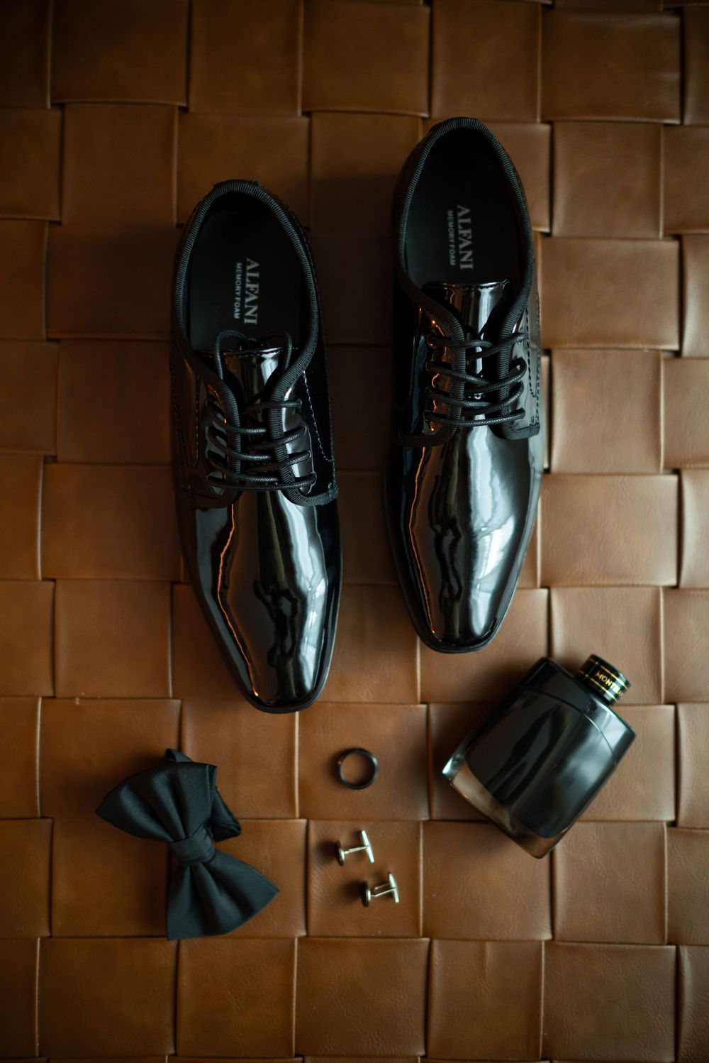 Groom's formal accessories including patent leather shoes, bow tie, and cufflinks on textured brown surface.