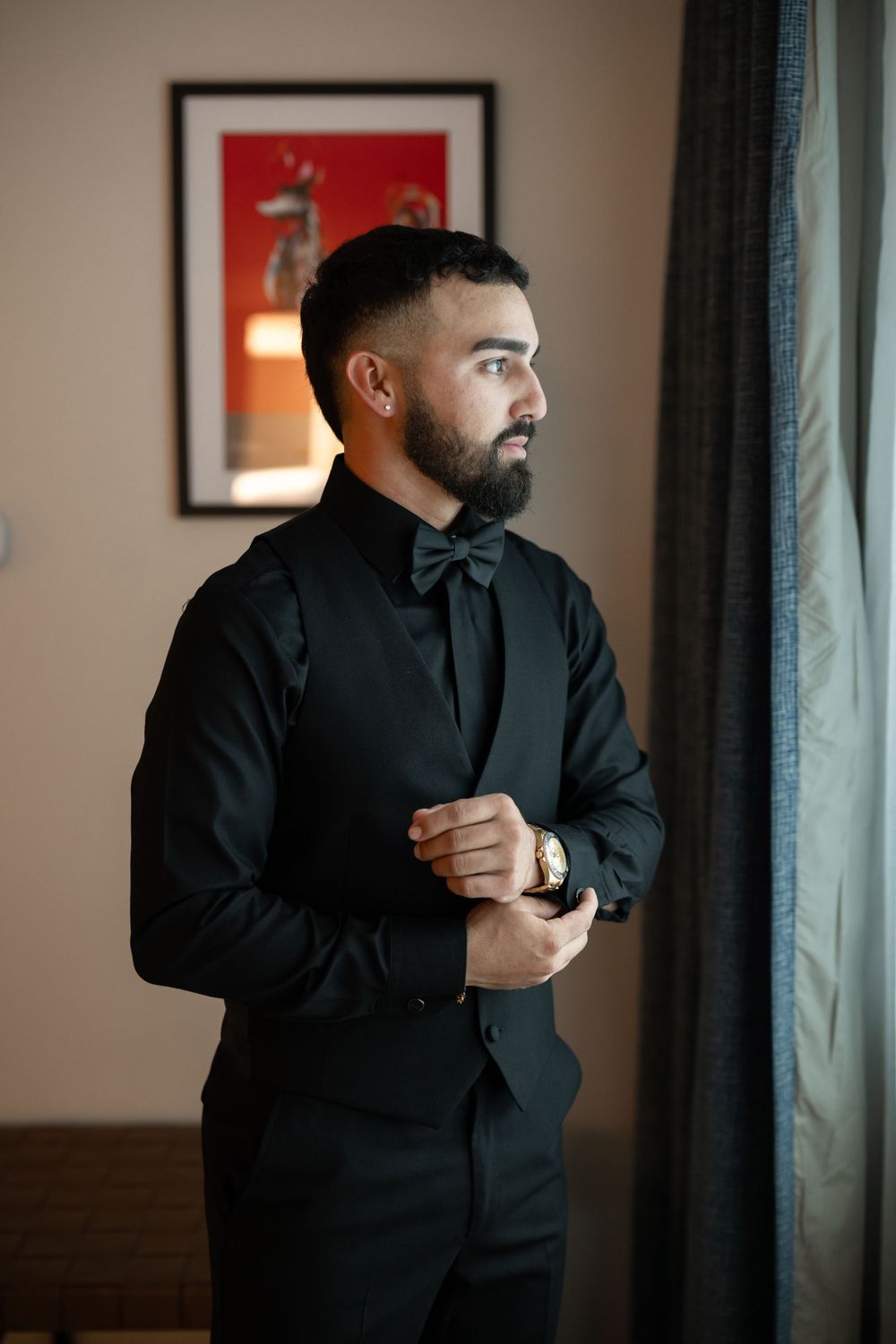 Elegant groom adjusts cufflinks while standing by window in formal black attire.