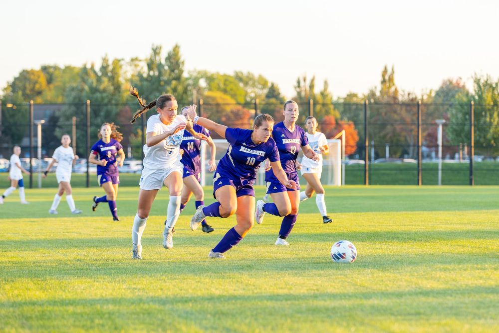 Soccer player makes a diving save attempt on grass field during competitive match in afternoon sunlight.